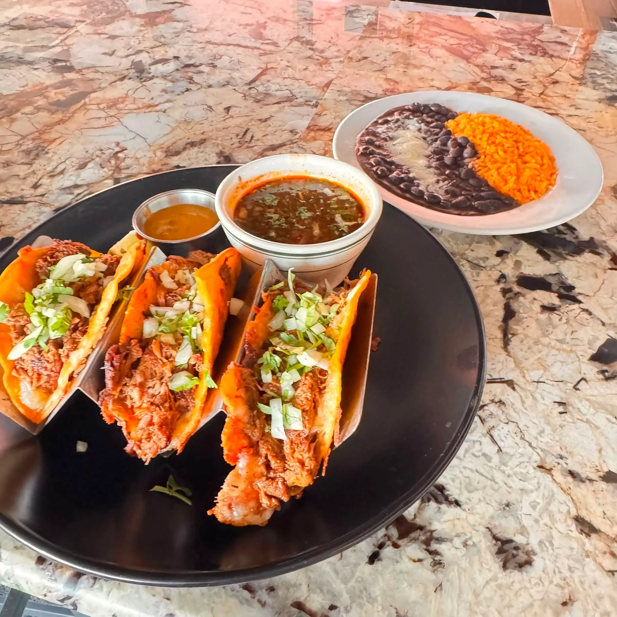 Birria tacos at Metztli — three crispy dorado tacos with braised beef and onion, served with a cup of consommé for dipping and a side of beans and rice