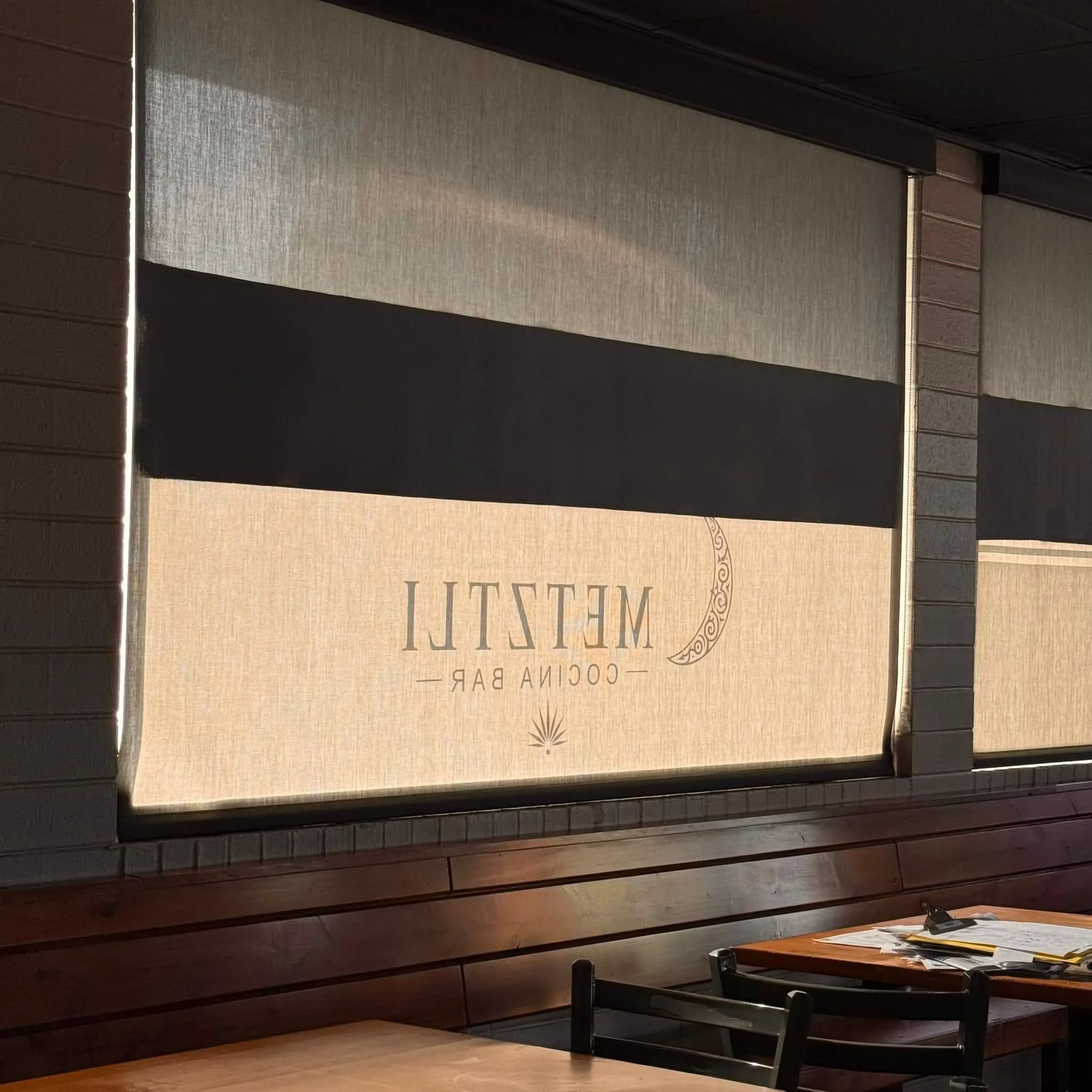 A window seat inside Metztli Cocina Bar with the Metztli crescent-moon logo printed on a linen shade, above wood-paneled banquette seating