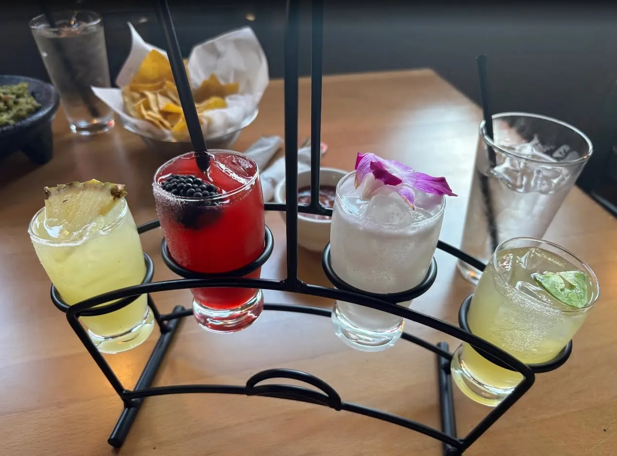 A tasting flight of four Metztli margaritas — pineapple, hibiscus-berry, coconut, and citrus — served on a curved iron caddy over a shared table