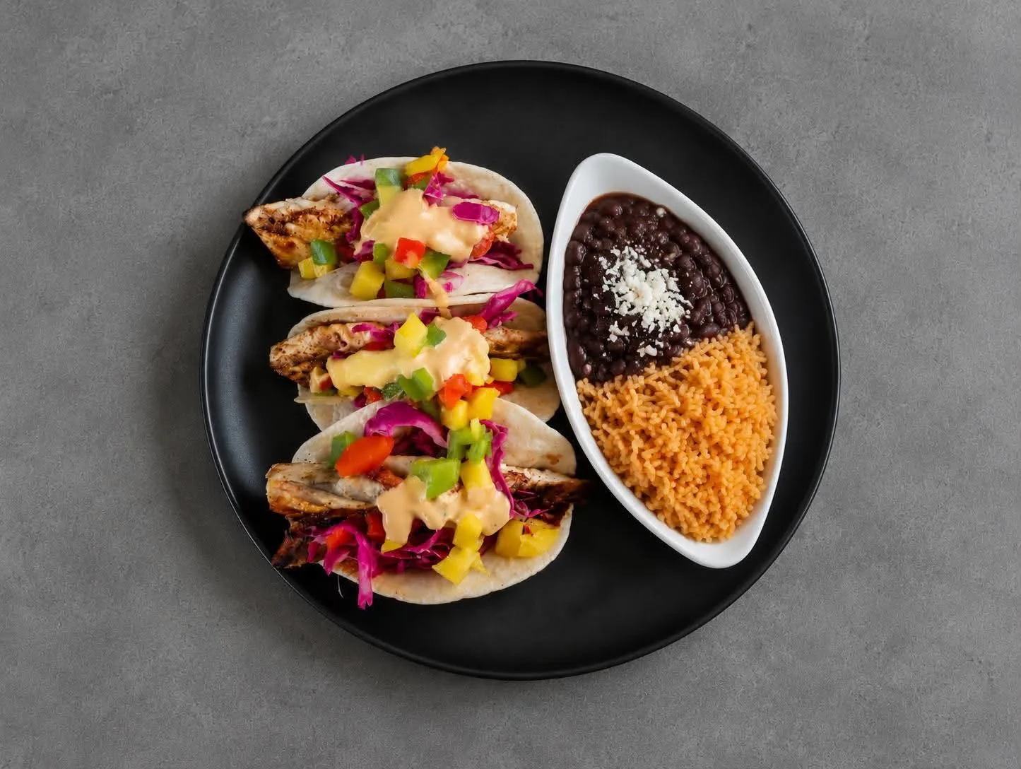Three grilled chicken tacos topped with mango-and-pepper salsa, red cabbage, and cream sauce, served on a black plate with rice and refried black beans